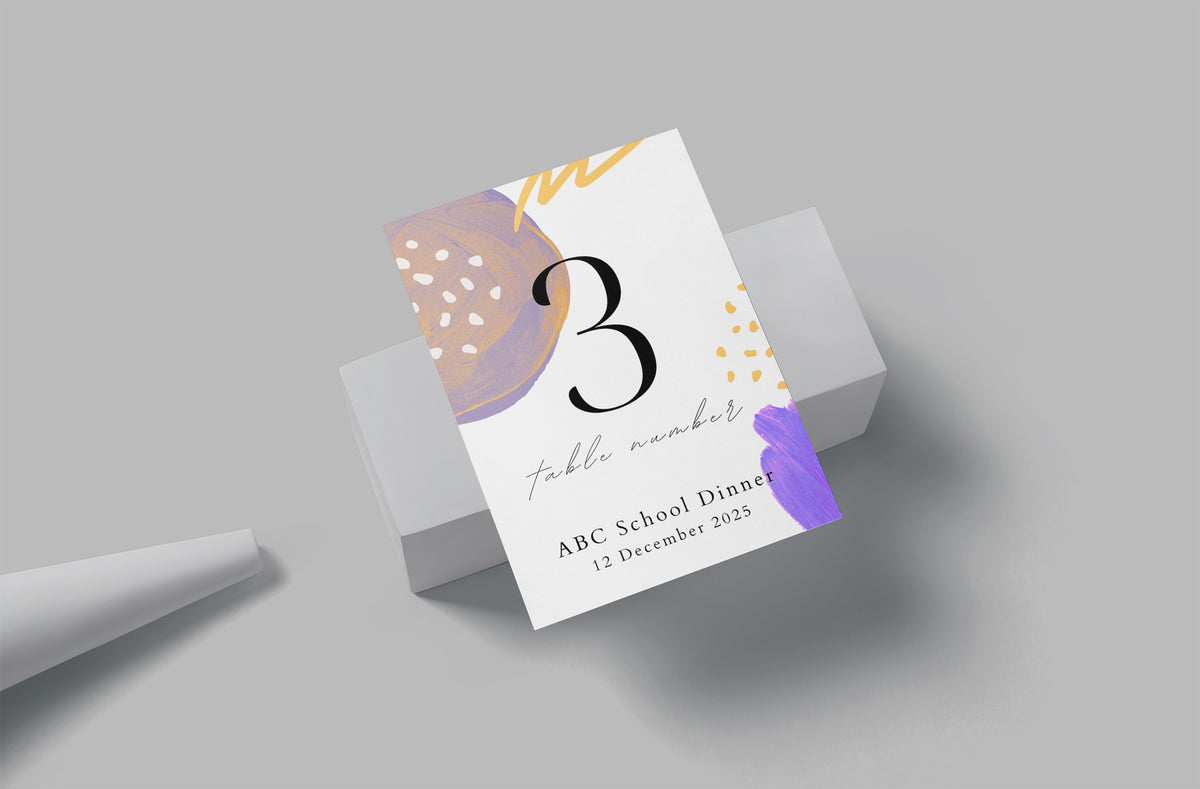 A printed table number card for an event, shown standing on a white pedestal against a light gray background. The card is white with abstract pastel artwork in purple, peach, and gold, and large text reading 'table number 3' and 'ABC School Dinner 12 December 2025'.