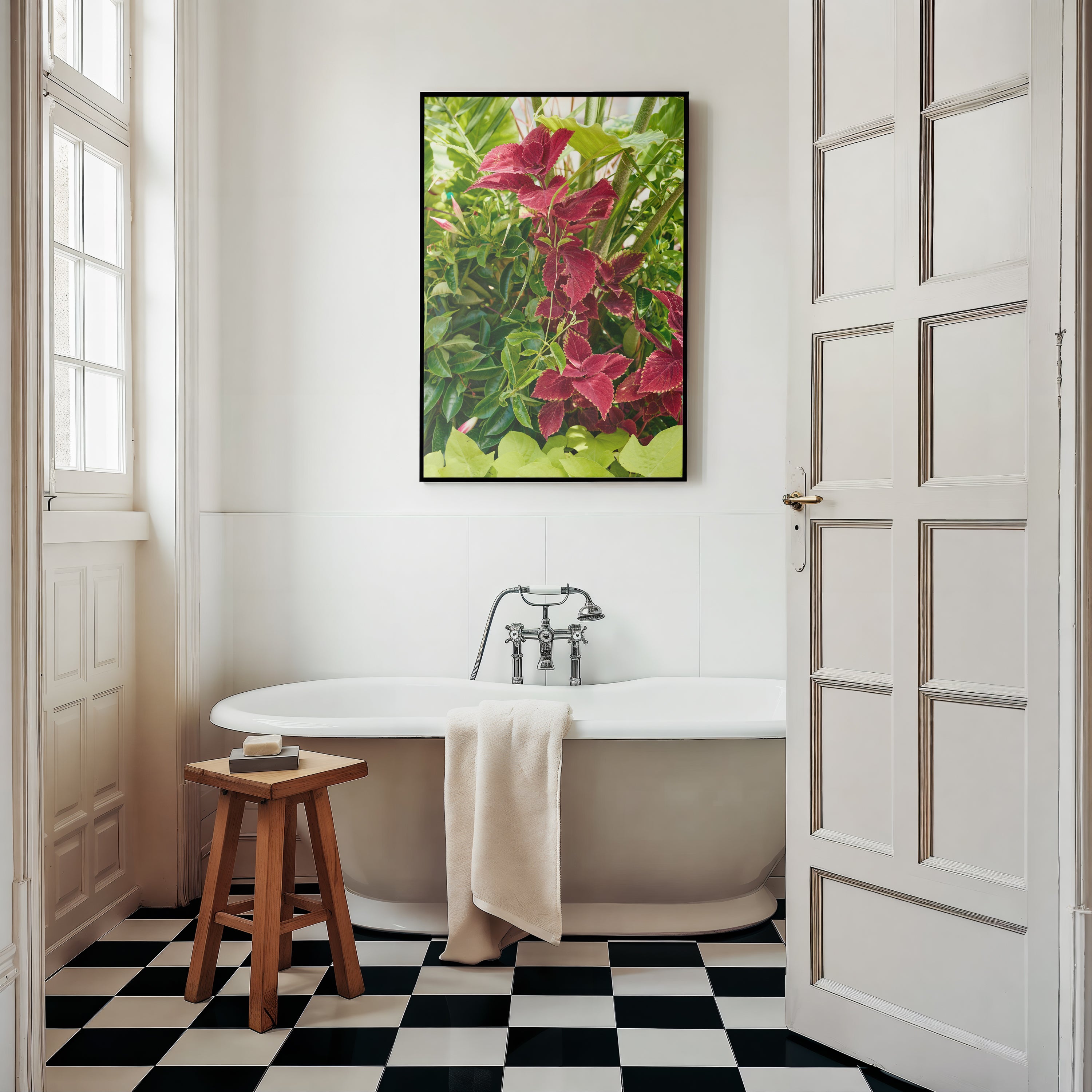 A framed wall art print featuring pink flowers and green leaves hangs above a white freestanding bathtub in a bright bathroom with black-and-white checkered flooring, a wooden stool, and white paneled doors.