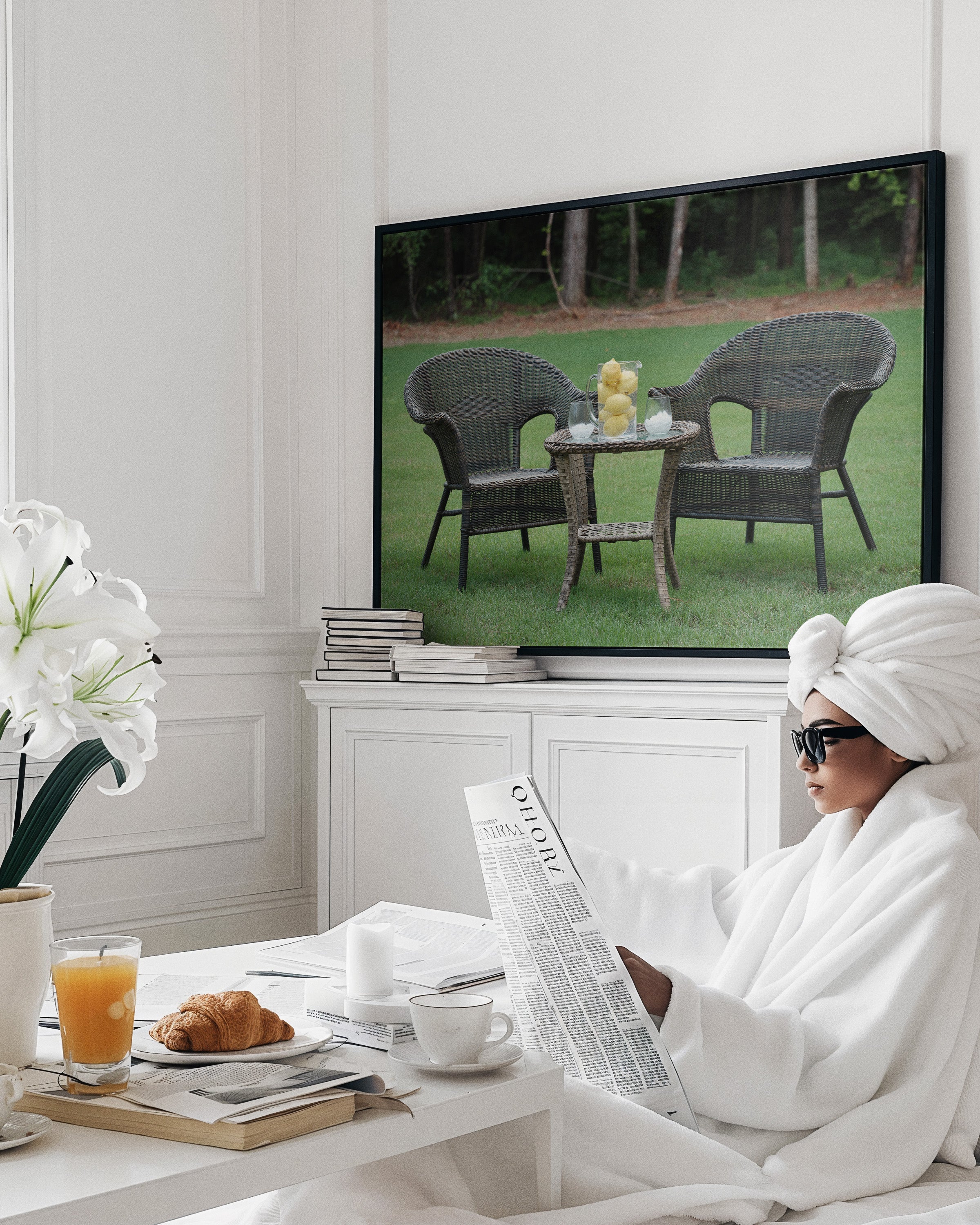 A framed wall art print hangs above a white cabinet in a bright, modern room. The print shows two wicker chairs and a small table on a green lawn.