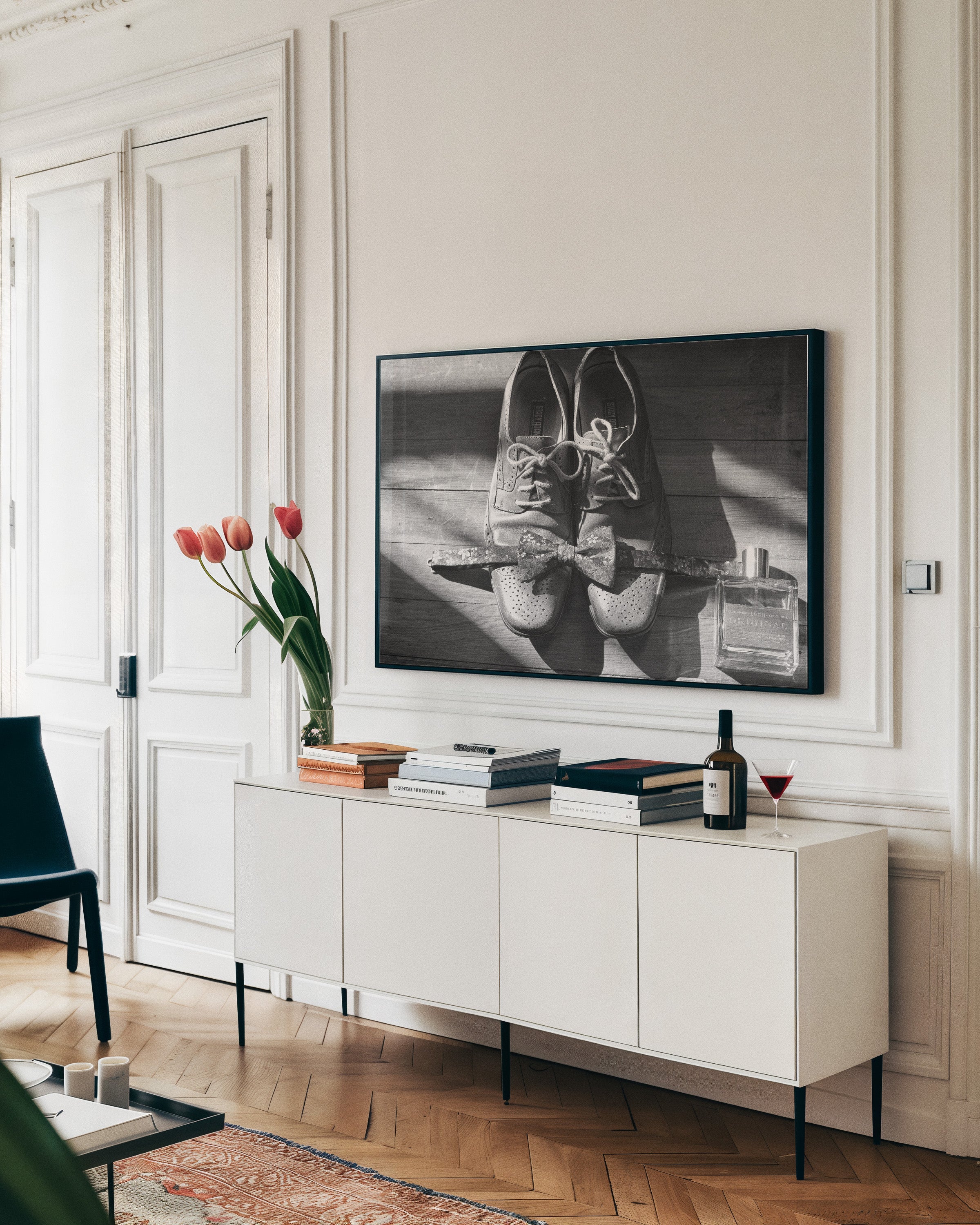 A framed black and white art print of a pair of lace-up shoes hangs on a white paneled wall above a white sideboard in a styled room with wood floors, books, and flowers.