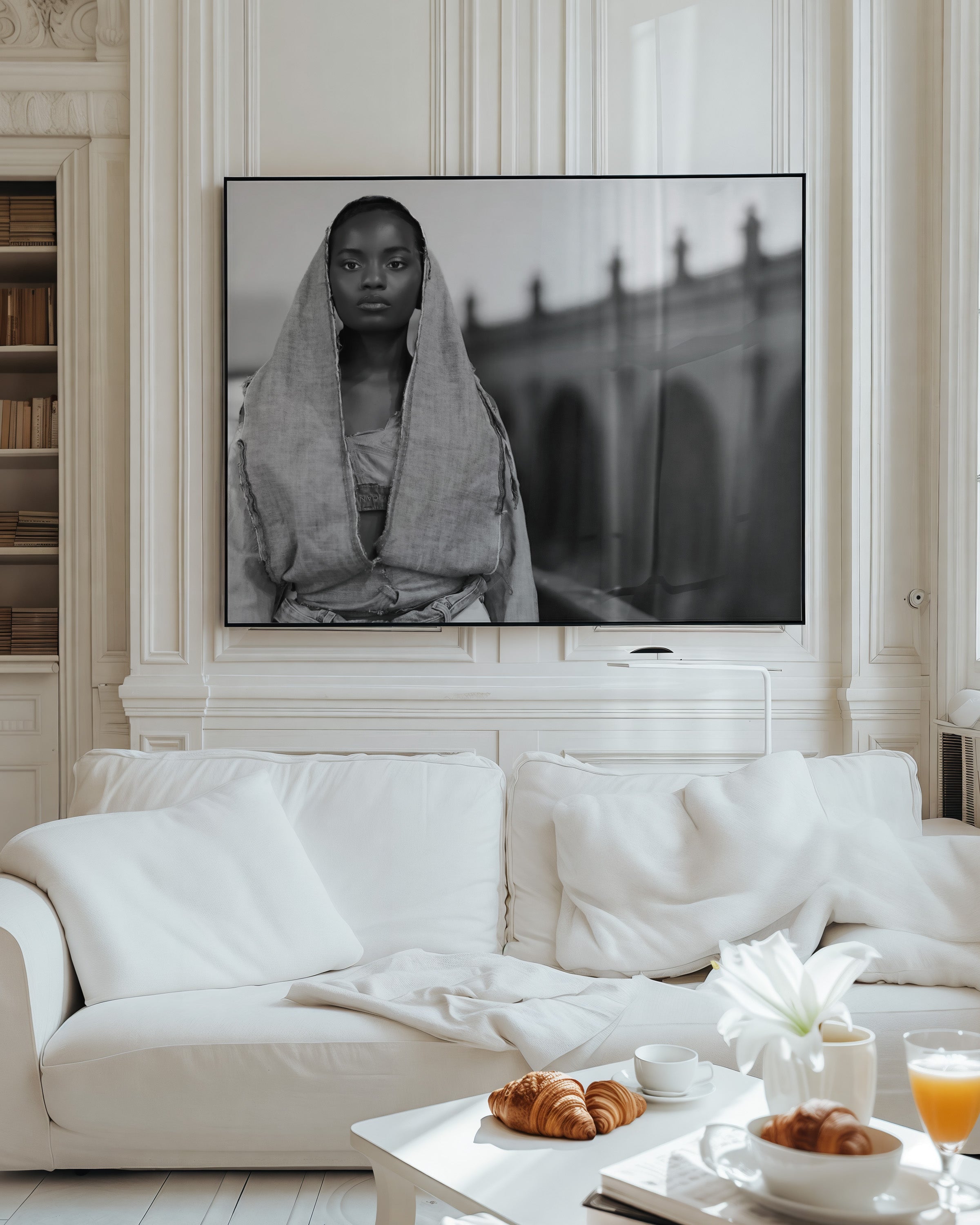 A large framed black and white art print of a woman draped in a shawl stands on a white wall above a white sofa in a bright, elegant living room.
