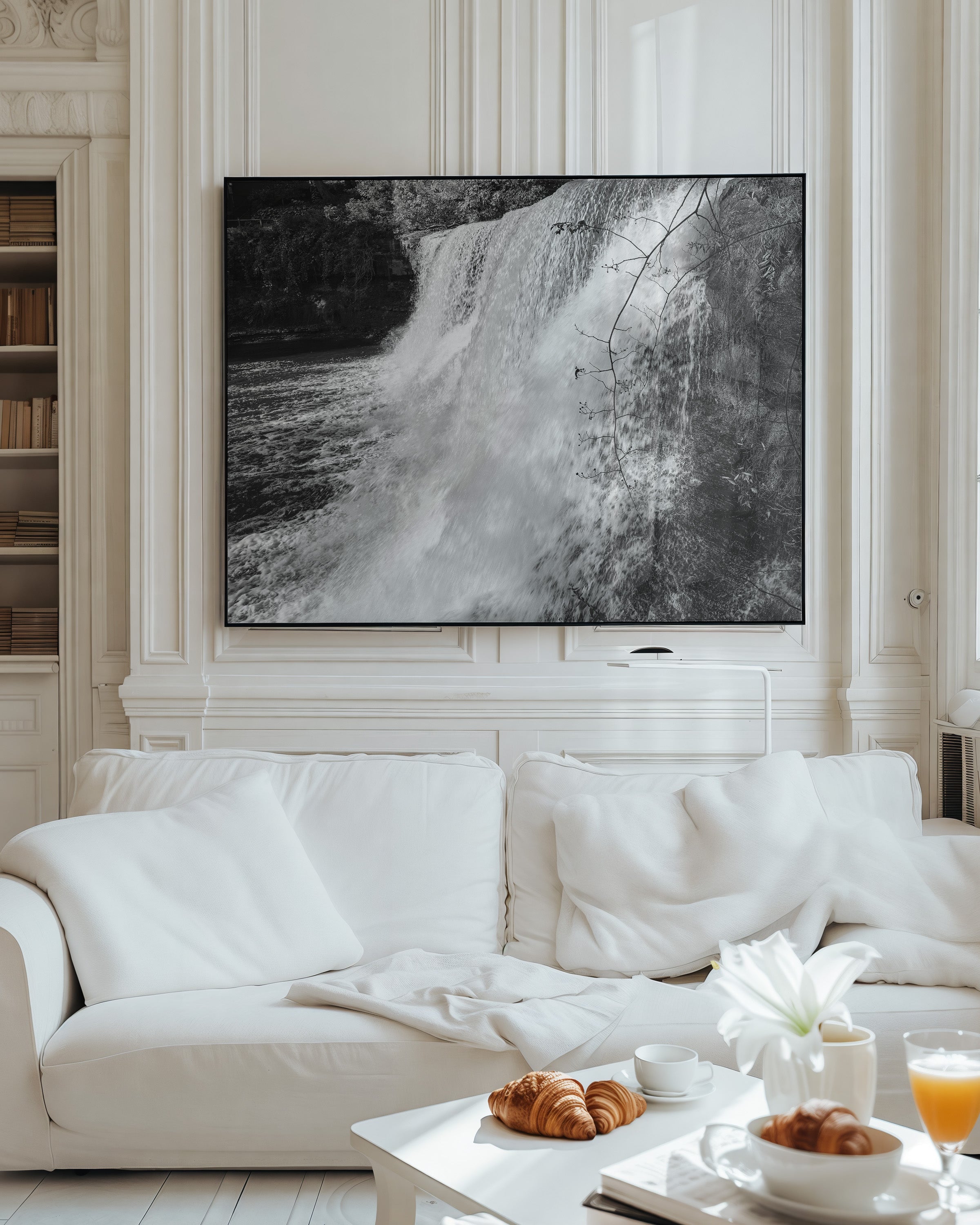 A large black and white framed wall art print depicting a waterfall, displayed in a classical white living room above a white sofa with a coffee table in the foreground.