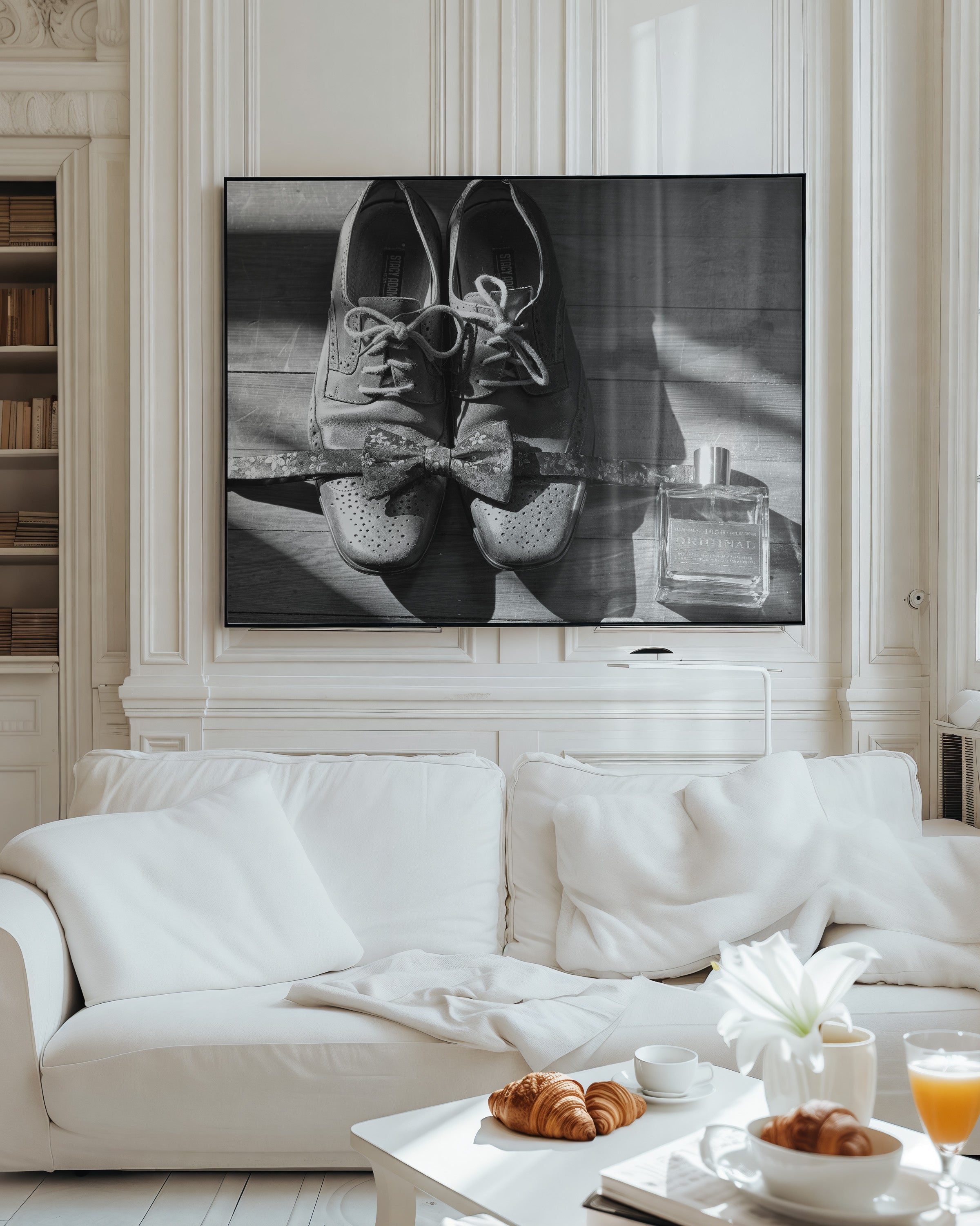 A large black and white framed wall art print depicting a pair of lace-up shoes with bows and a perfume bottle, displayed above a white sofa in a bright, elegant living room.