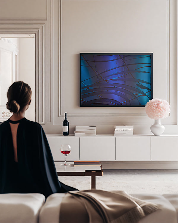 A woman sits in a modern living room with her back to the camera, facing a framed abstract blue and black wall art piece above a white media console. The room has elegant cream walls, a low coffee table with a glass of red wine, and a vase with a pink pom-pom decoration.