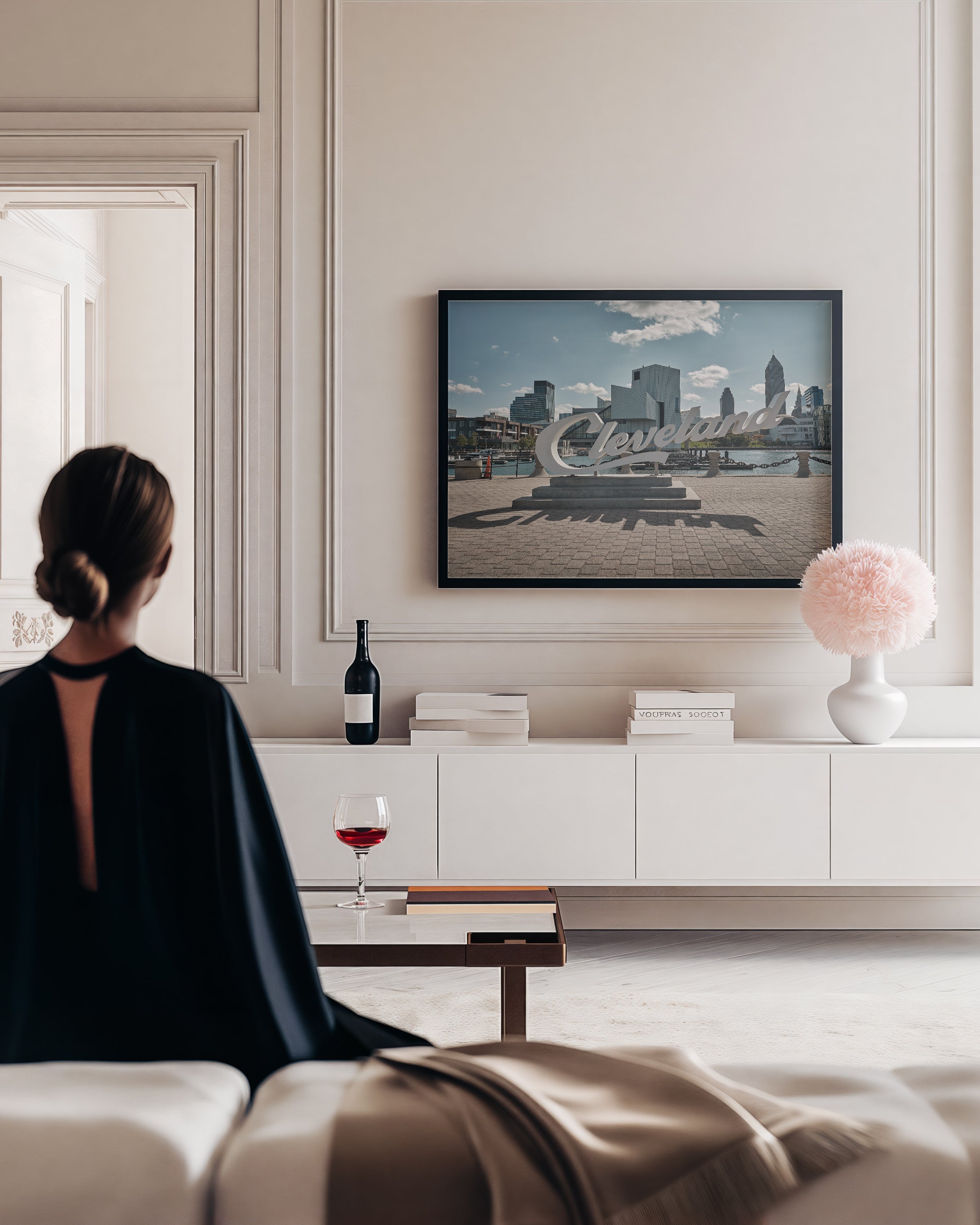 A framed art print of the Cincinnati skyline with a large 'Cincinnati' script overlay, displayed on a cream wall in a modern living room. A woman in a black backless top sits on a sofa in the foreground, looking at the screen.