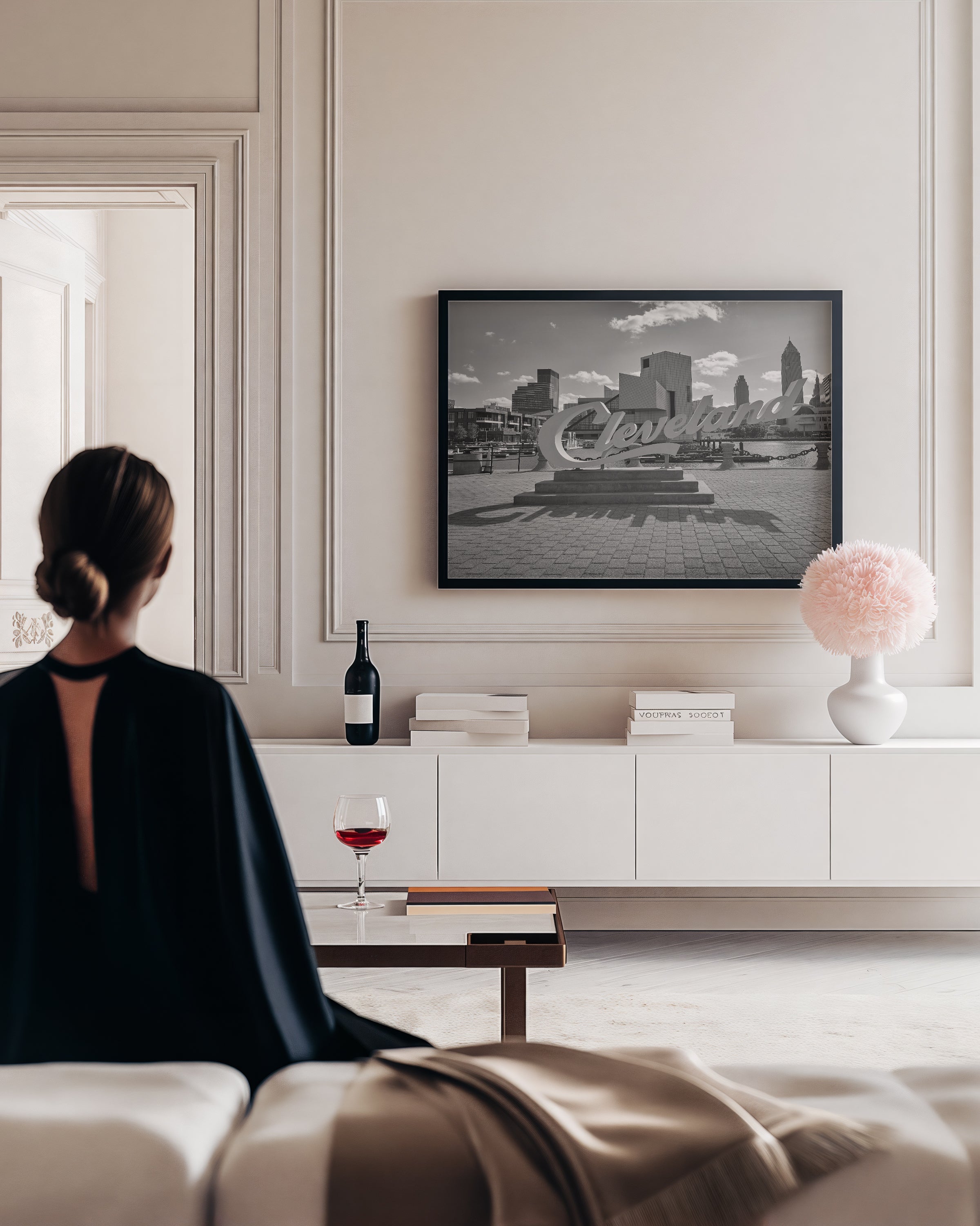 A black and white framed art print of the Cleveland skyline is displayed on a cream-colored wall in a modern living room setting, viewed from behind a woman sitting on a sofa.