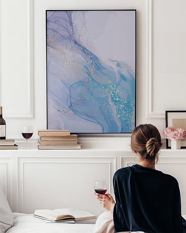 A woman sits on a white sofa in a bright living room, holding a glass of red wine and facing a large framed abstract wall art print. The artwork features flowing blue and lavender-purple patterns with scattered gold glitter-like speckles.