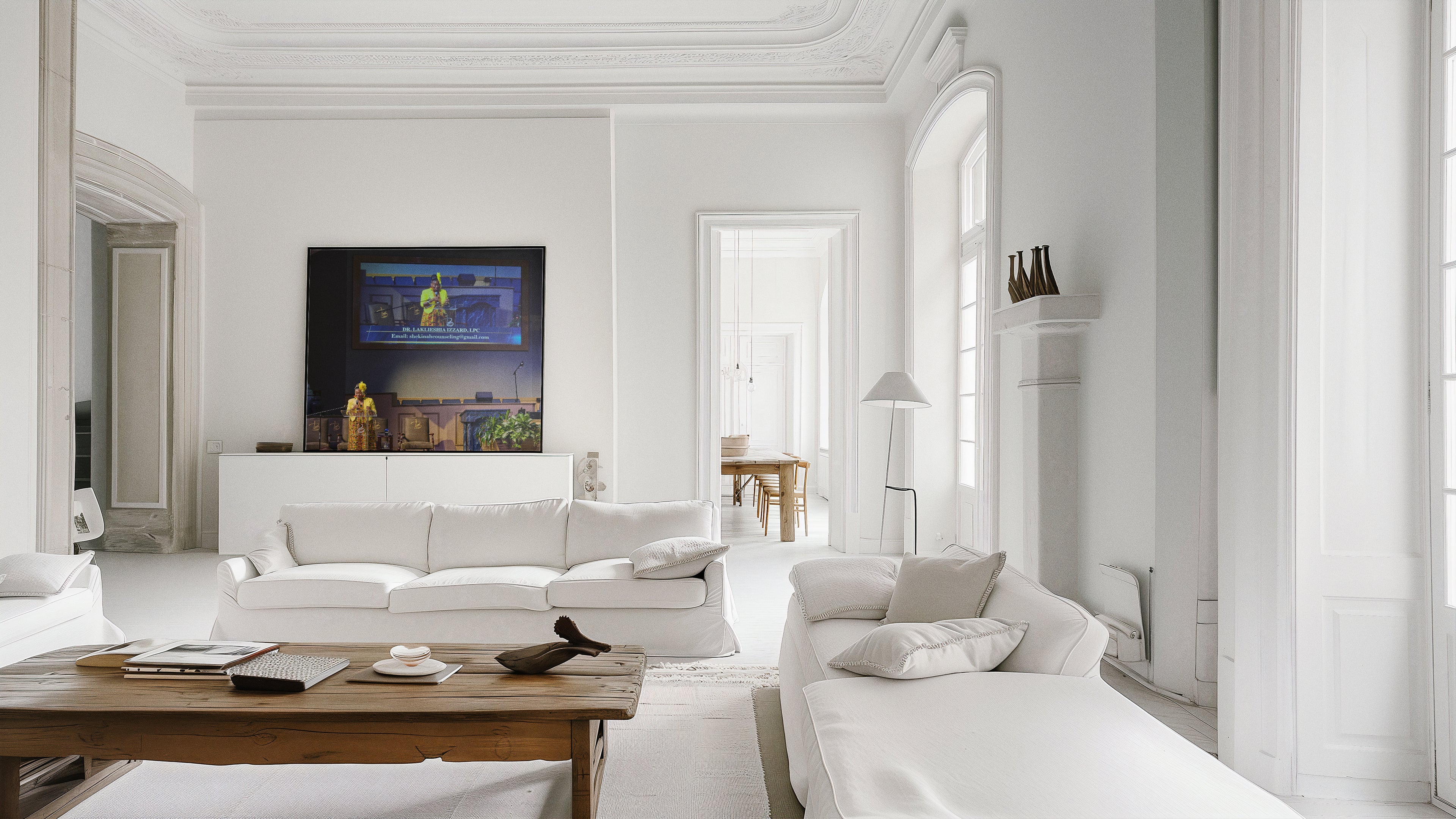 A bright, modern living room with white walls and furniture, featuring a white sofa, a wooden coffee table, and a large framed artwork on the wall.