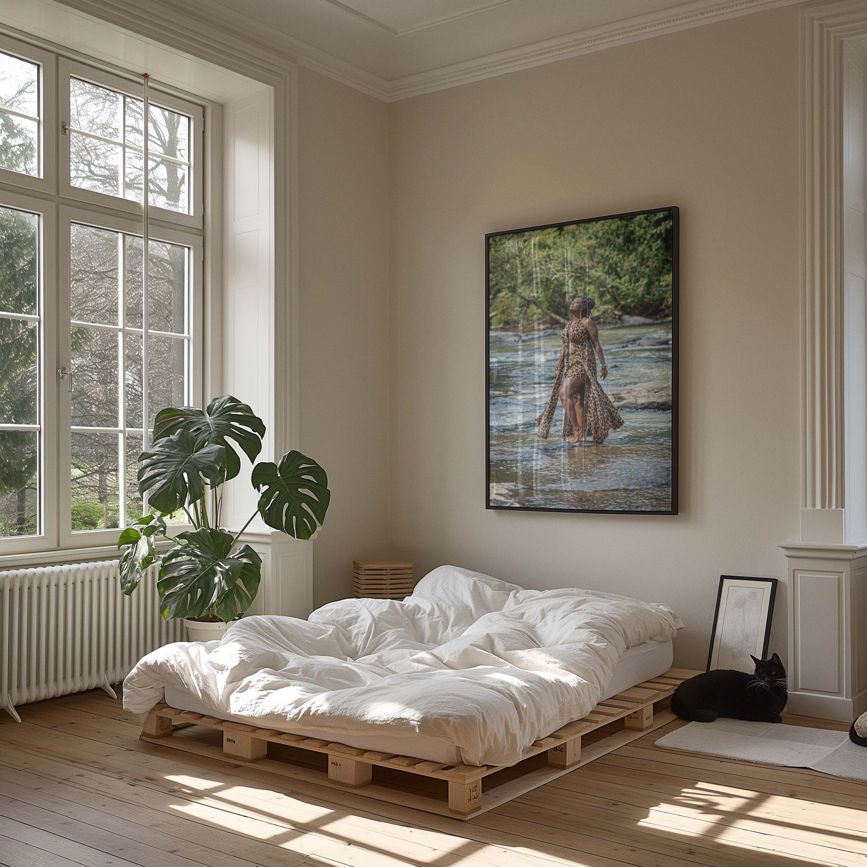 A framed vertical art print depicting a woman standing in a river, displayed on a beige wall in a modern, sunlit minimalist bedroom with a wooden bed and a black cat.