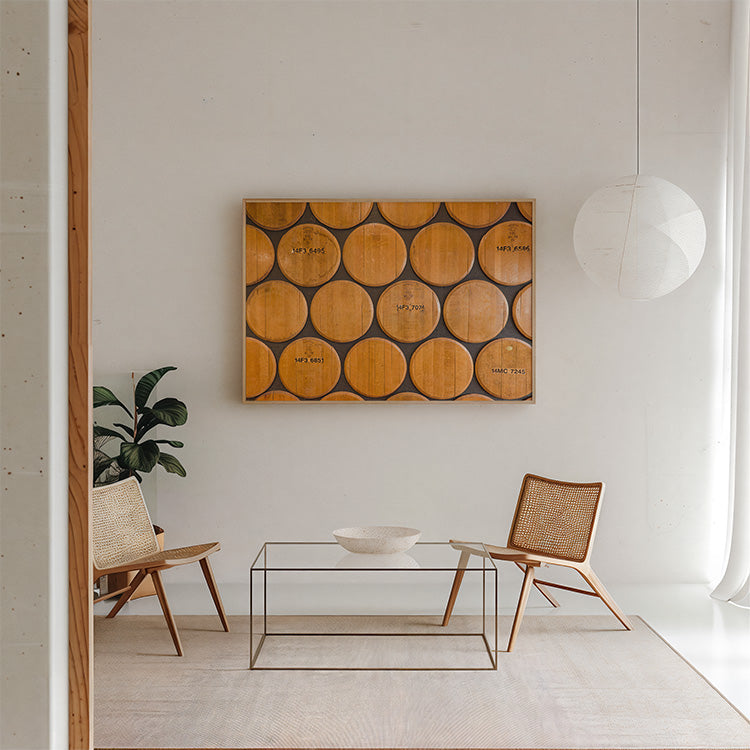 A rectangular wall art piece made of stacked wooden wine barrels hangs on a white wall in a modern living room setting with two woven-back chairs, a glass coffee table, and a hanging pendant light.