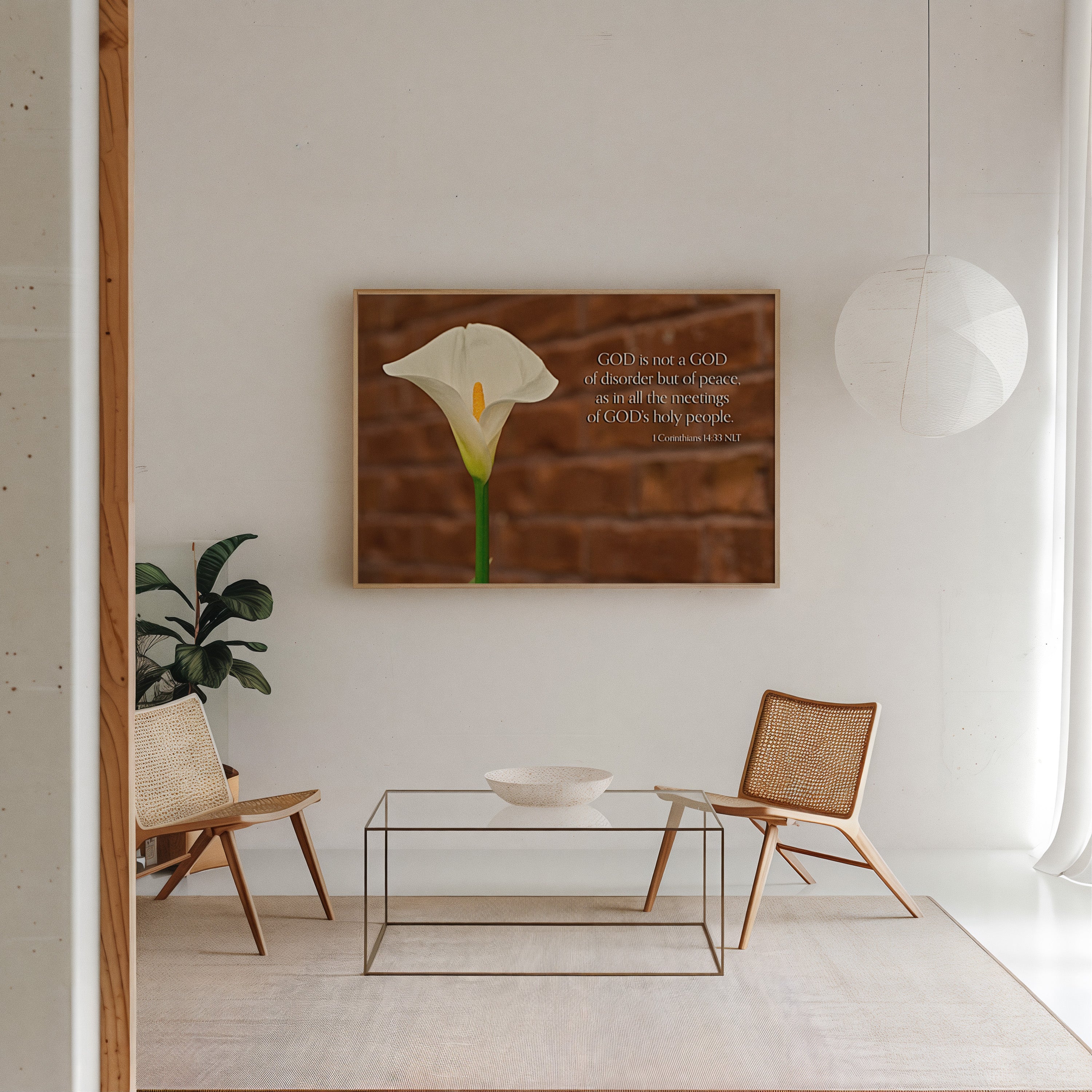 A framed wall art print featuring a white calla lily on a brown wood-textured background with a Bible verse, displayed in a bright, modern living room setting with two woven chairs and a glass coffee table.