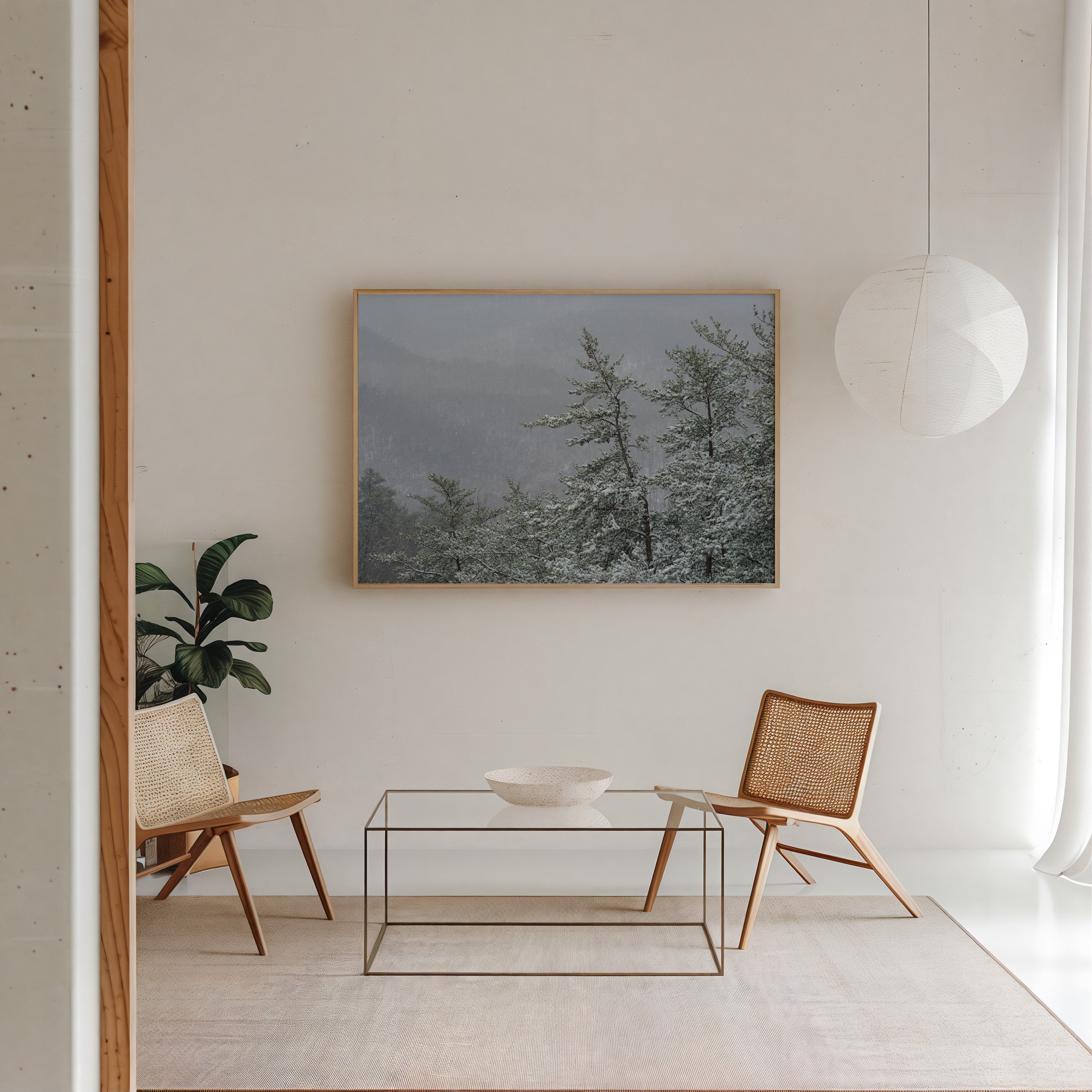 A framed landscape art print depicting snow-covered pine trees under a misty sky, displayed on a white wall in a modern, minimalist living room setting with two wooden chairs and a glass coffee table.