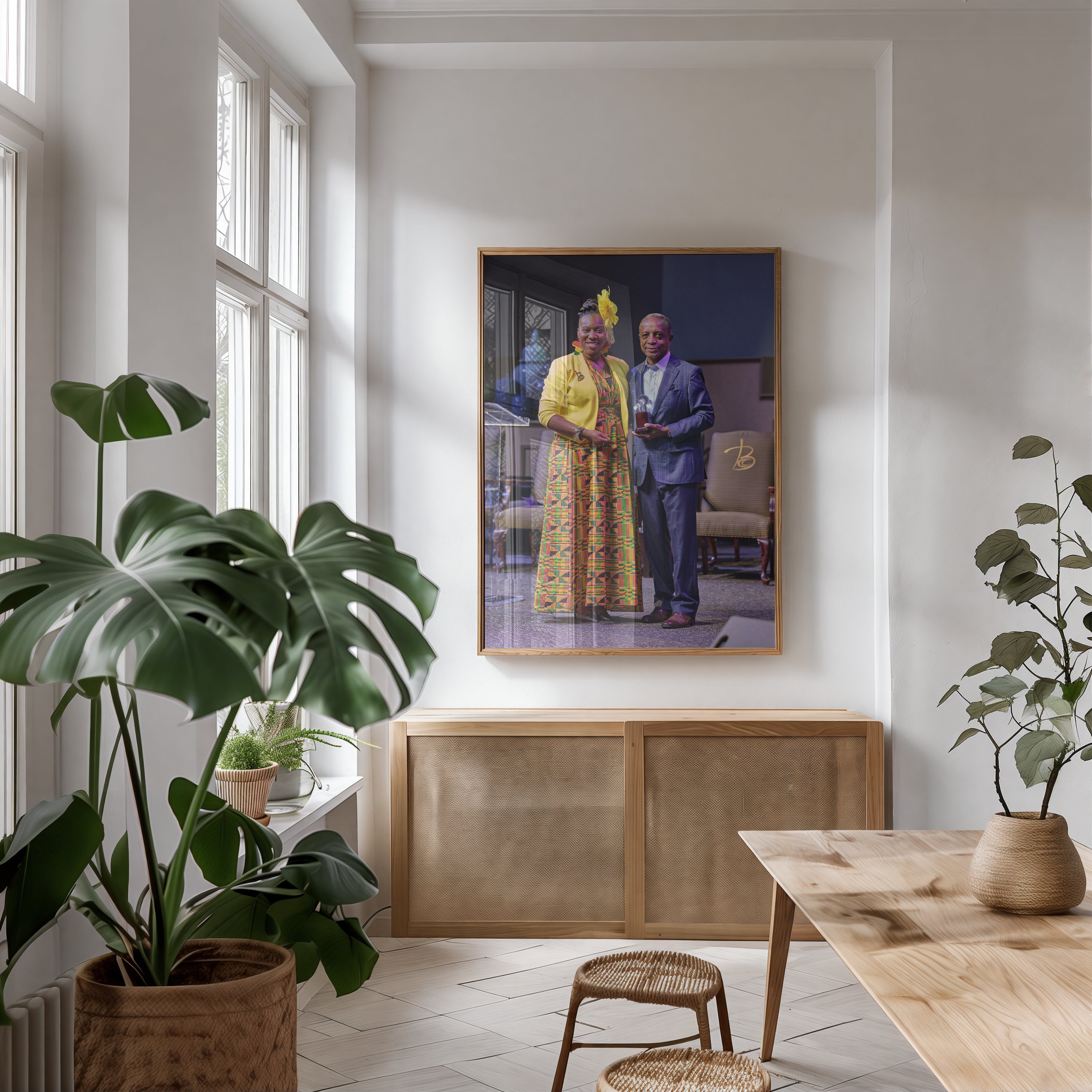 A framed portrait print hangs on a white wall above a light wood cabinet in a bright, modern room with large potted plants and a wooden table.