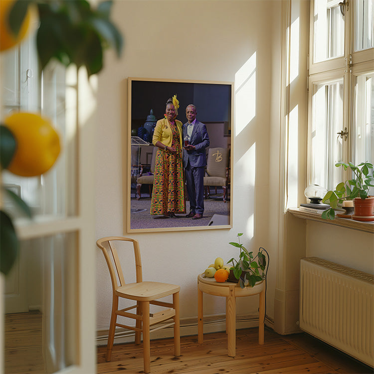 A framed vertical portrait print of a man and woman standing indoors, hung on a cream wall in a sunlit room with a wooden chair, small table, plants, and windows.
