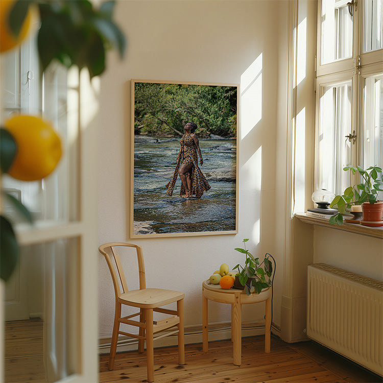 A framed art print depicting a woman standing in a shallow river surrounded by lush green trees, displayed on a cream wall in a bright, modern room with a wooden chair, side table, and potted plants.