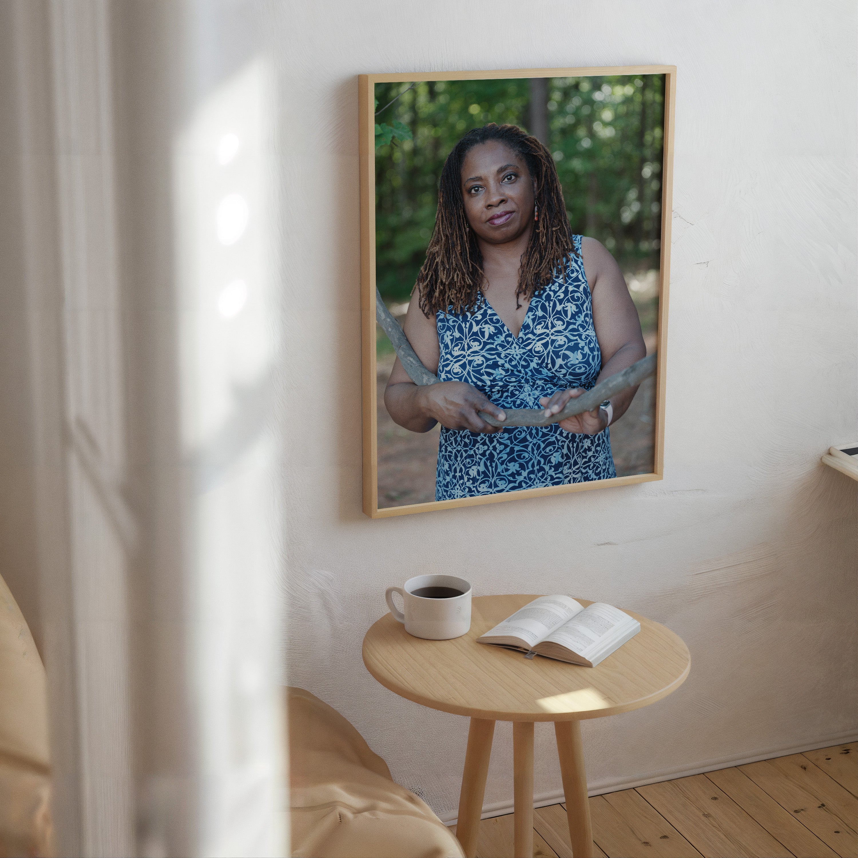 A framed vertical portrait print of a woman in a blue patterned top hanging on a light wall above a small round wooden table with a mug and an open book.