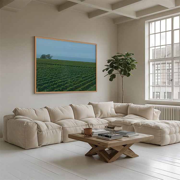 A large framed landscape art print of a green field under a blue sky hangs on a beige wall above a cream tufted sectional sofa in a bright, modern living room.