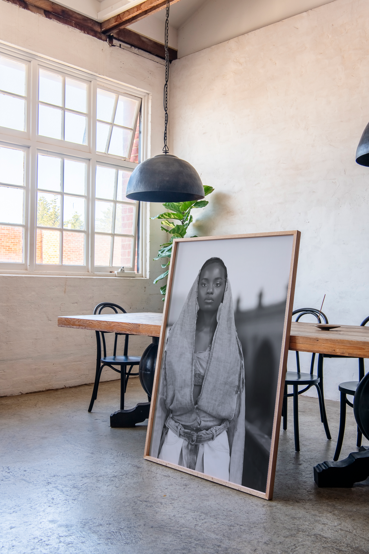 A large framed black and white portrait print of a person in traditional clothing leans against a wooden table in a bright, modern loft-style room with concrete floors, a large window, and a hanging pendant light.