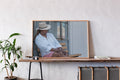 A large framed art print with a light wood frame hangs on a textured white wall above a wooden console table. The print features a photograph of a woman wearing a white shirt, pink skirt, and a beige fedora hat, sitting on a wooden chair.