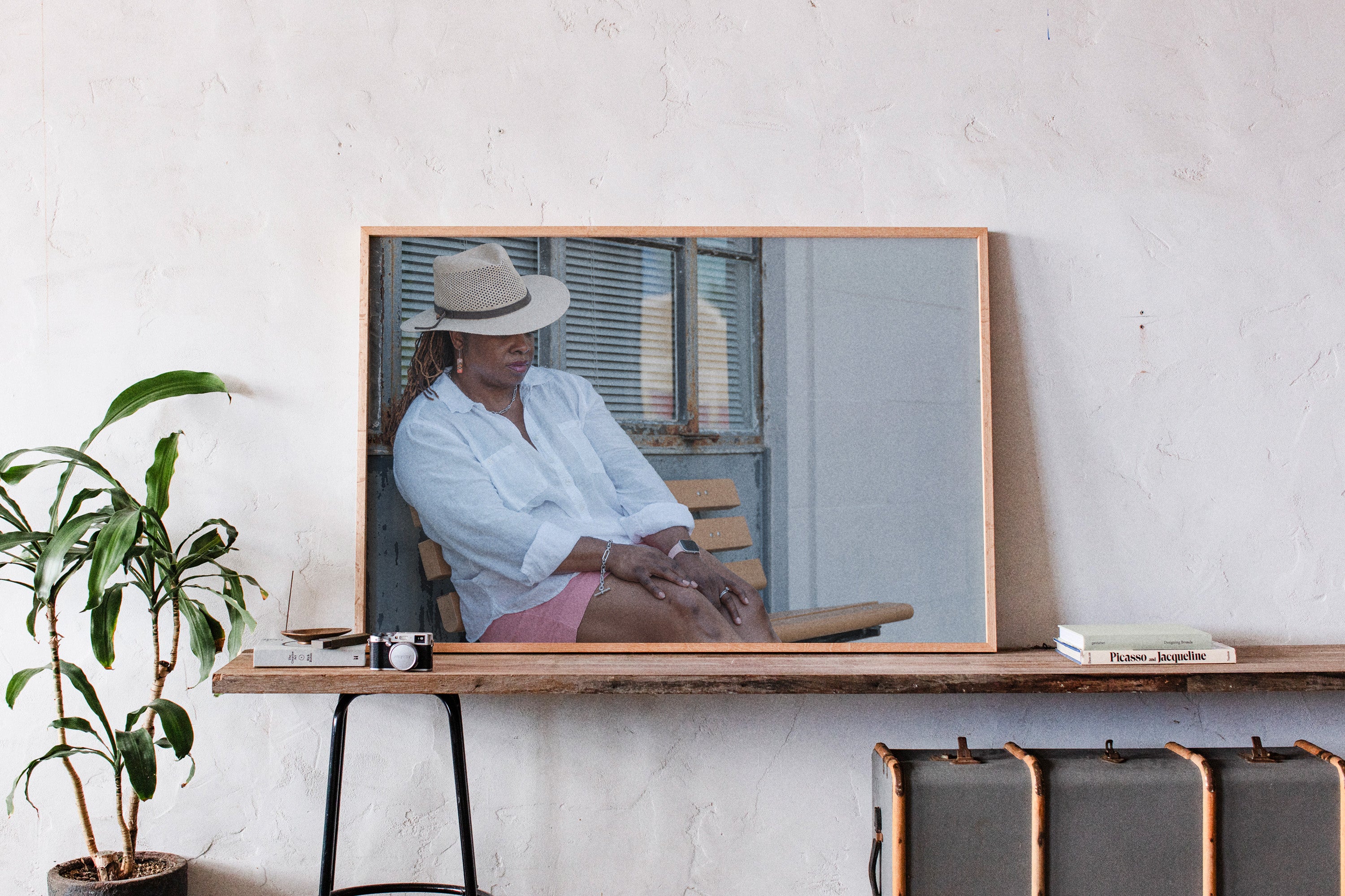A large framed art print with a light wood frame hangs on a textured white wall above a wooden console table. The print features a photograph of a woman wearing a white shirt, pink skirt, and a beige fedora hat, sitting on a wooden chair.