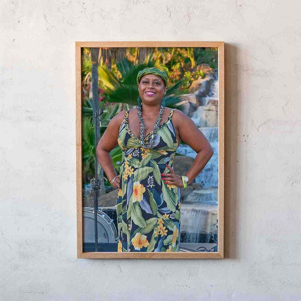 A framed vertical portrait print of a smiling woman in a tropical floral dress and headwrap, mounted on a light wood frame and hanging on a textured off-white wall.