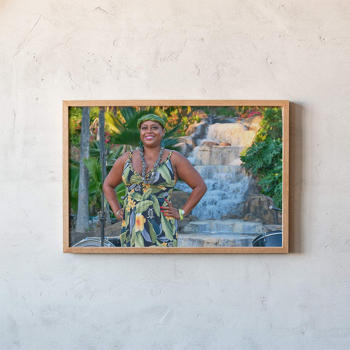 A framed photographic print of a smiling woman in a green floral dress standing in front of a waterfall, mounted on a textured off-white plaster wall.