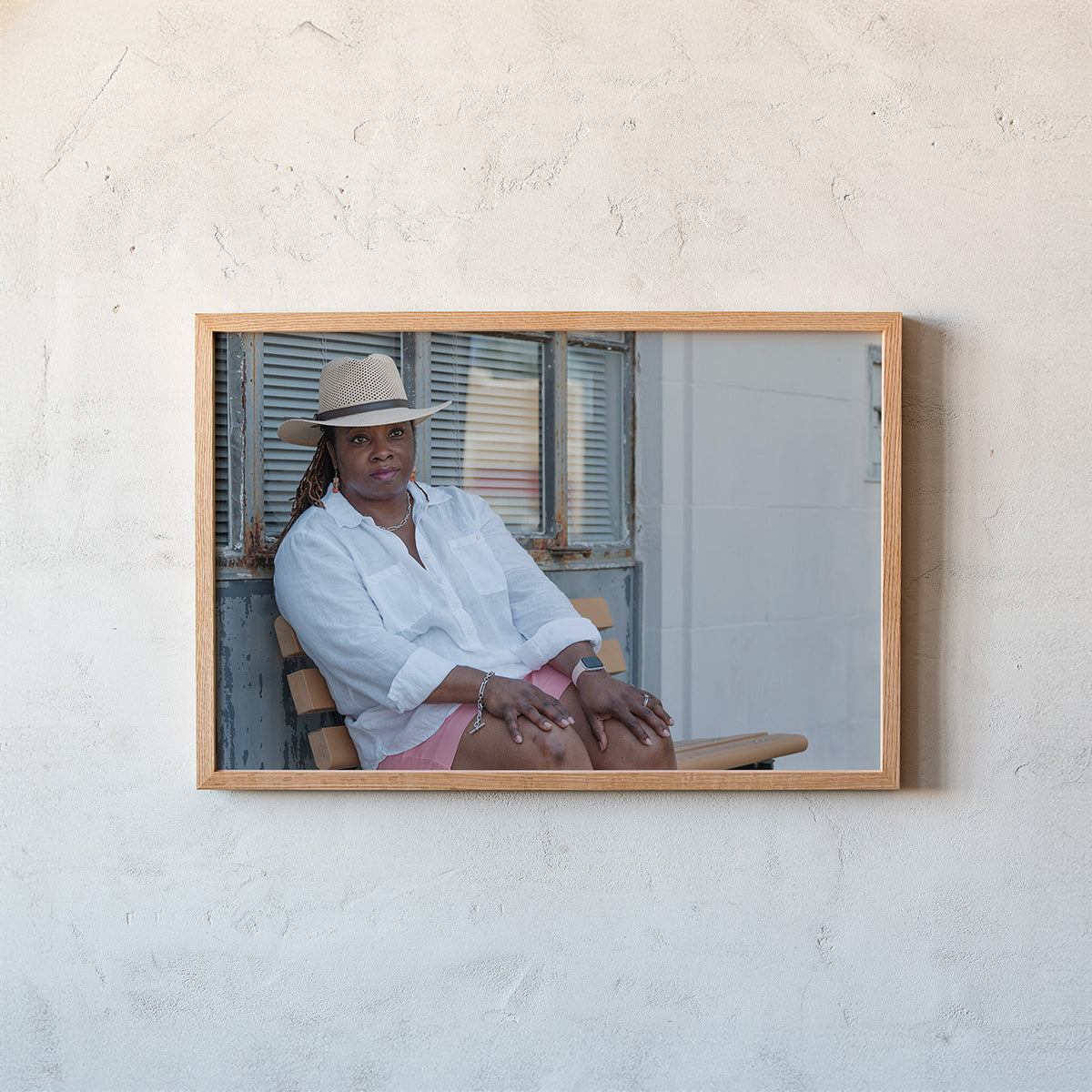 A framed photographic print of a woman in a white shirt and hat sitting on a wooden chair, mounted on a textured off-white plaster wall.