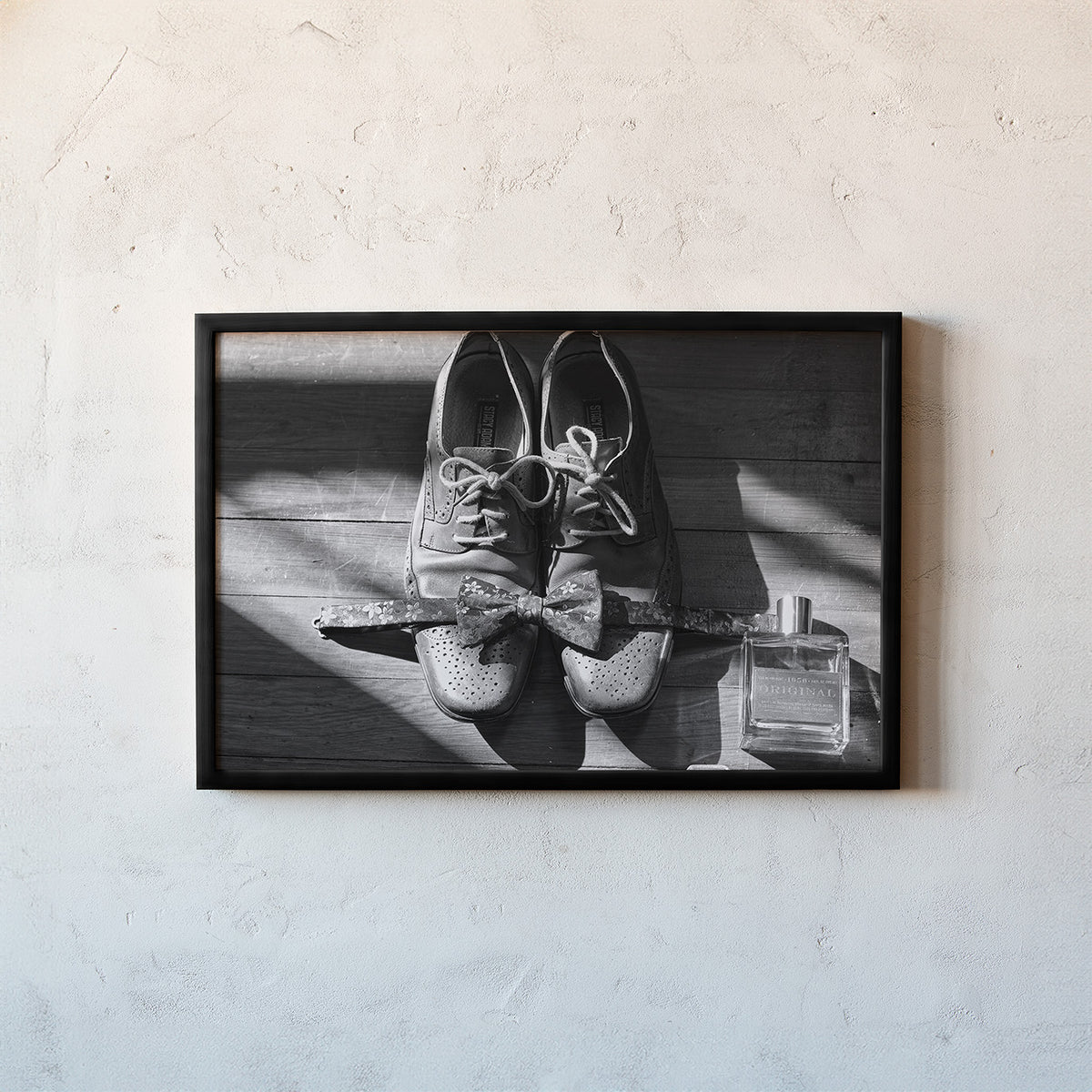 A framed black and white photograph hanging on a textured light gray wall. The photo inside the frame shows a pair of lace-up shoes tied with a bow, with a perfume bottle nearby.