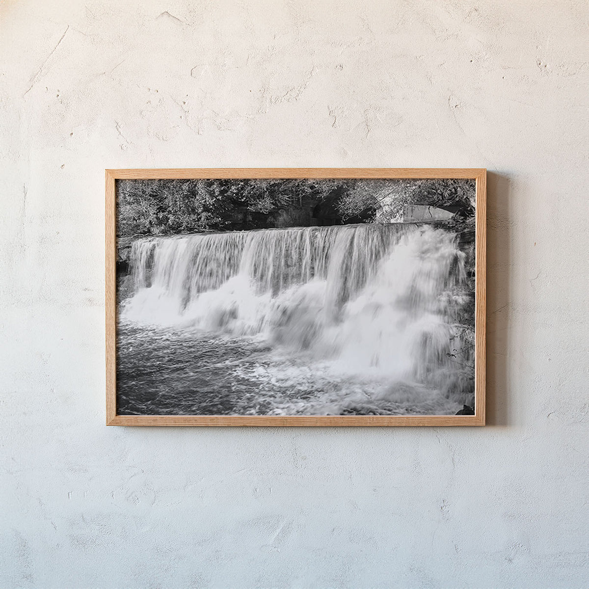 A framed black and white landscape photograph of a waterfall, presented in a light natural wood frame and mounted on a textured off-white plaster wall.