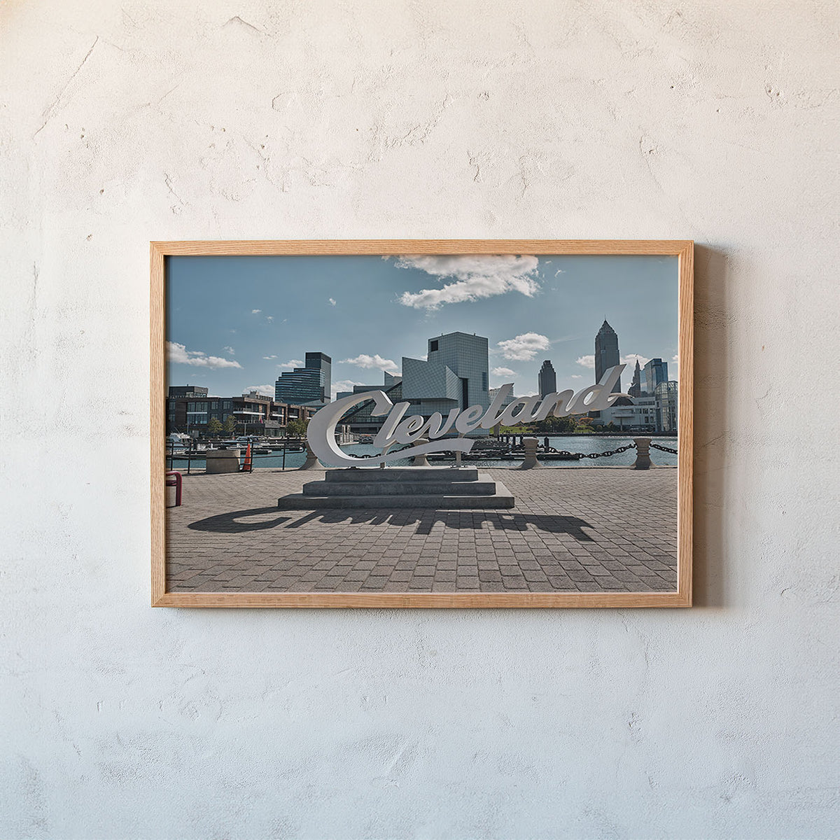 A framed photographic print of the Cleveland skyline and the large 'Cleveland' sculpture in the foreground, mounted on a textured off-white wall.