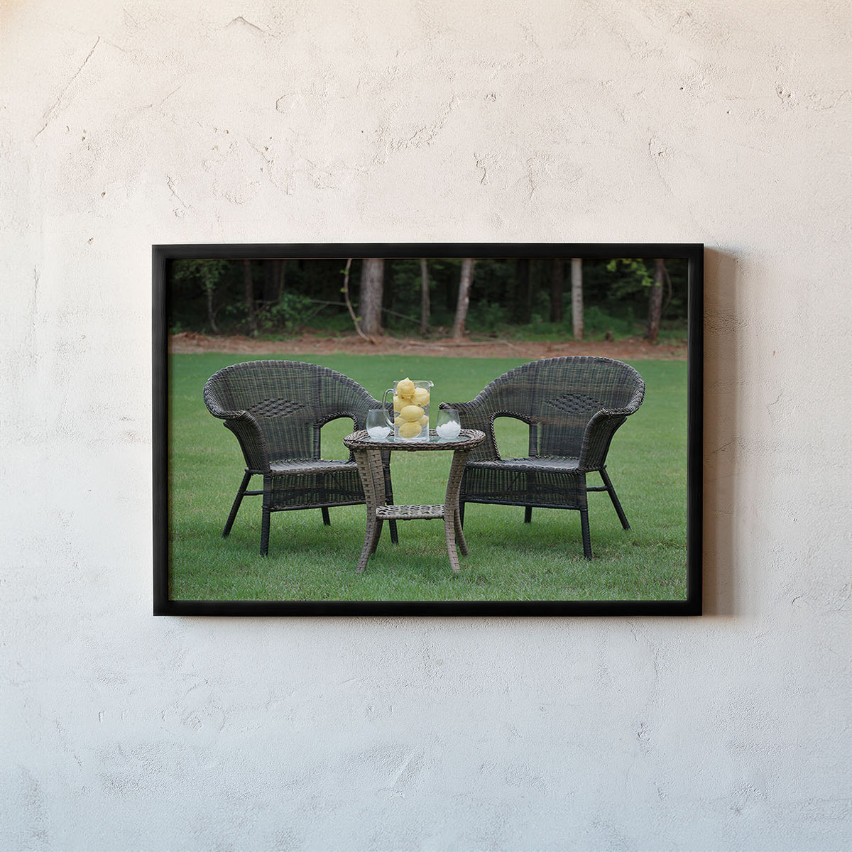 A framed photographic print of a wicker patio set on a green lawn, displayed on a textured off-white wall.