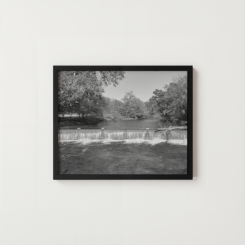 A black-framed black and white photographic print of a small waterfall in a park, displayed on a light gray wall.
