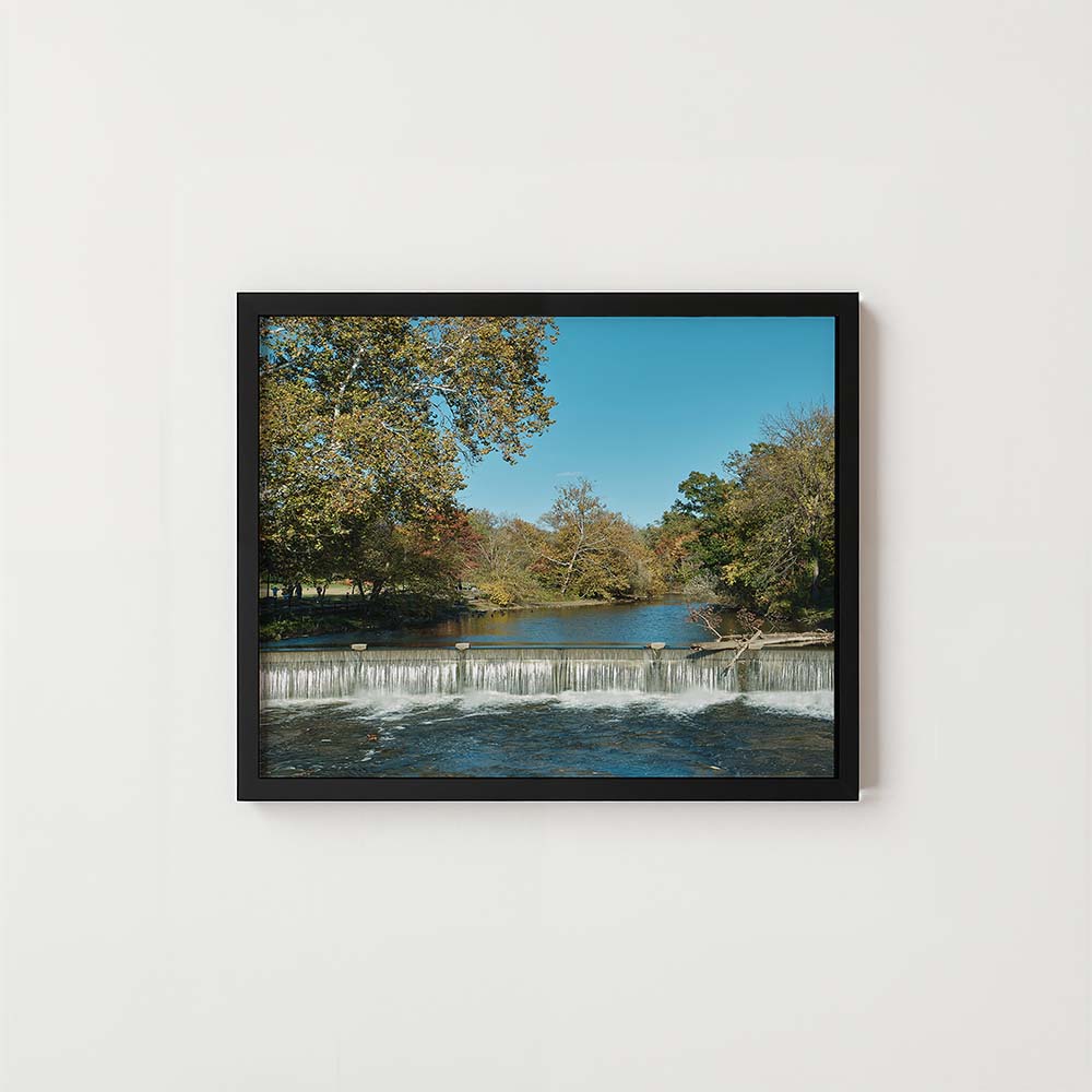 A framed landscape photograph of a small waterfall over a river, surrounded by autumn trees and a blue sky, displayed on a clean white wall.