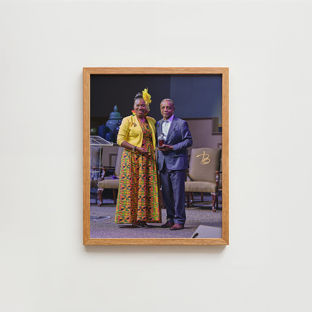 A framed photographic art print hangs on a light gray wall. The print shows a man in a blue suit and a woman in a yellow patterned dress with a yellow hat, standing indoors.