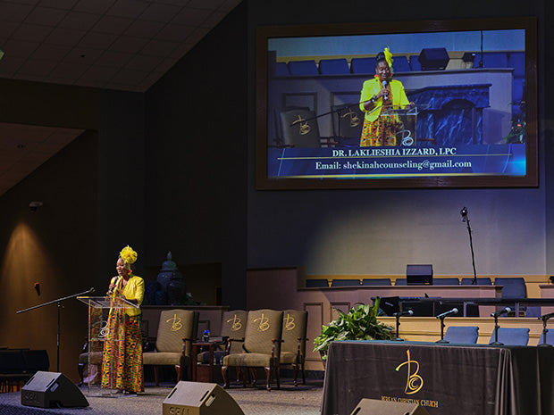 A woman in a bright yellow and patterned outfit stands at a podium on a church stage, with a large screen behind her showing a live feed of the same speaker. The stage includes chairs, a table with a draped front, and speakers.