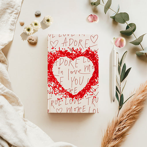 A squarefold journal with a red glittery heart design and handwritten red text on a cream cover, styled in a flat lay with flowers, greenery, a pen, and neutral textiles on a light beige background.