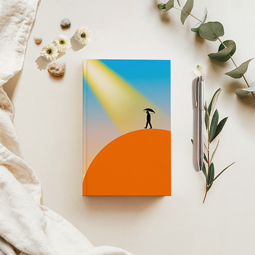 A hardcover notebook with a minimalist cover illustration of a person walking on an orange hill under a blue sky with a yellow spotlight, styled in a flat lay with a pen, eucalyptus leaves, small flowers, and a textured cream fabric on a light beige background.