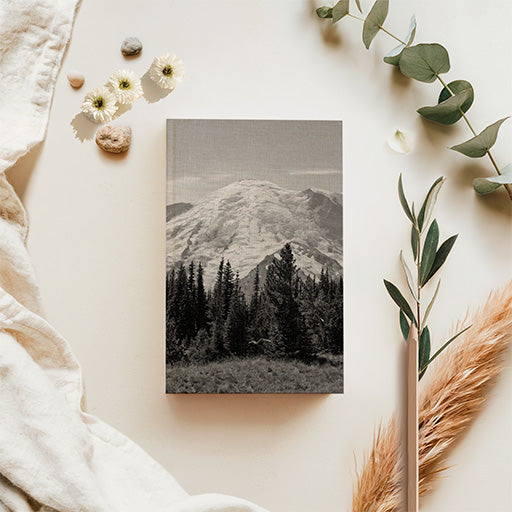 Mountain Majesty SquareFold journal featuring black-and-white photography of snow-covered Mount Rainier above evergreen forests, displayed with botanical accessories.