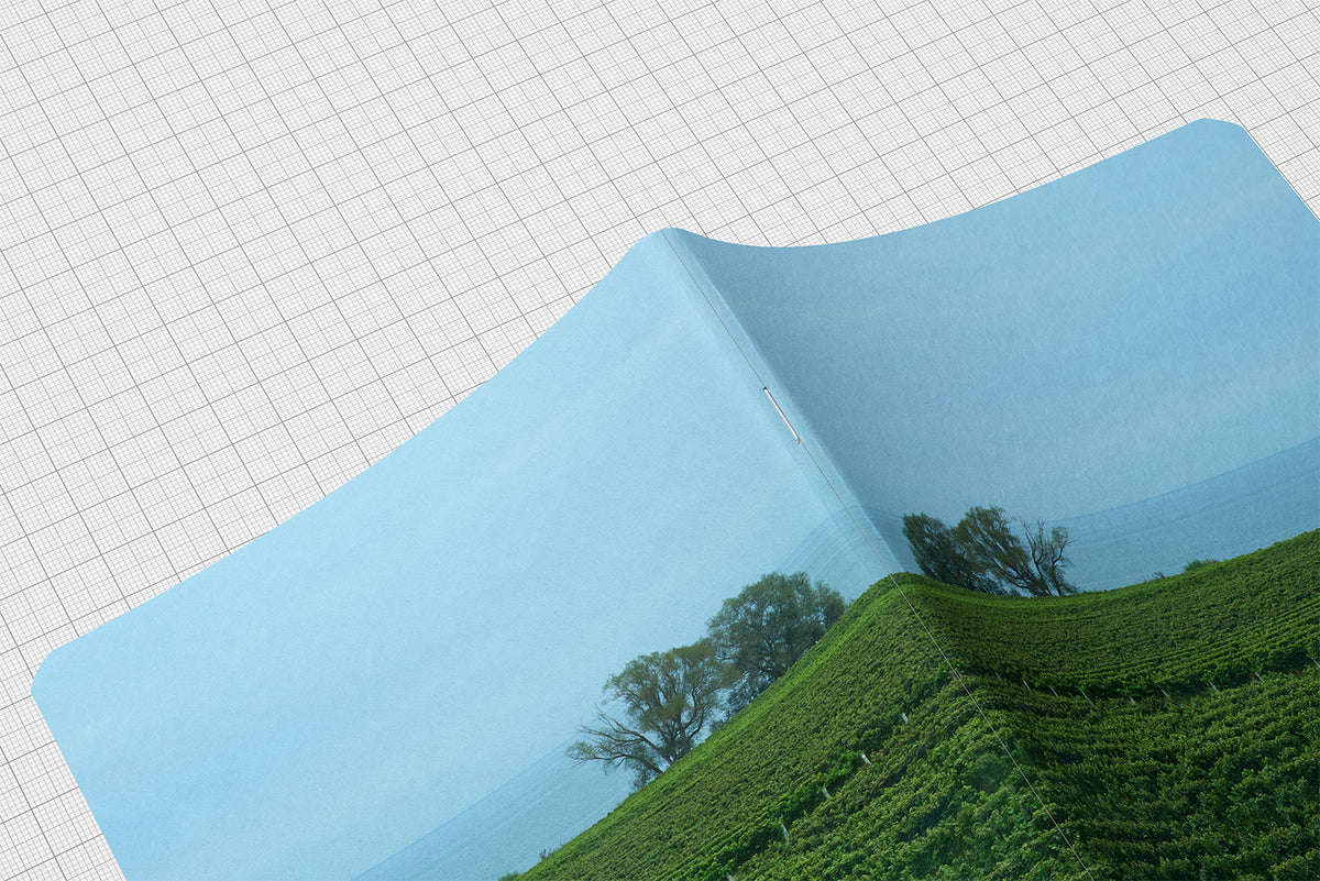 Vineyard Artist Series journal featuring rows of lush green grapevines extending toward a distant horizon beneath a blue sky. Shown partially peeled back to reveal a printed landscape scene of green vineyard hills and trees under a blue sky. The sheet is laid diagonally on a white grid background.