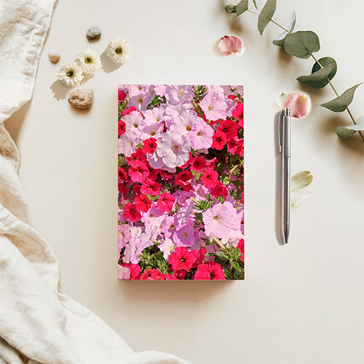 A floral journal with a vibrant print of pink, red, and purple flowers, styled in a flat-lay arrangement on a light beige surface with a silver pen, eucalyptus leaves, and small stones.