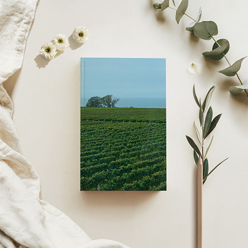 A squarefold journl featuring a scenic photograph of a green vineyard under a blue sky, styled in a flat lay with a cream linen cloth, white flowers, and eucalyptus branches on a warm beige background.