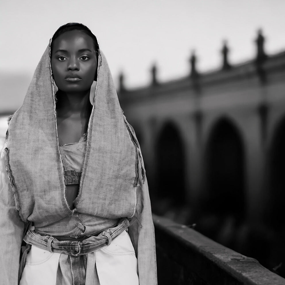 A black and white portrait of a woman wearing a draped linen-textured hooded garment over her head and shoulders, standing on a bridge with blurred arches in the background.