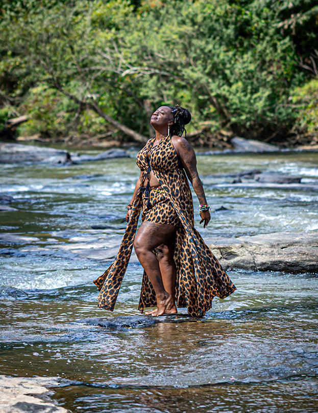 A woman stands barefoot in a shallow river wearing a leopard-print two-piece outfit with a halter top and matching skirt or shorts. Trees and rocks are visible in the background.