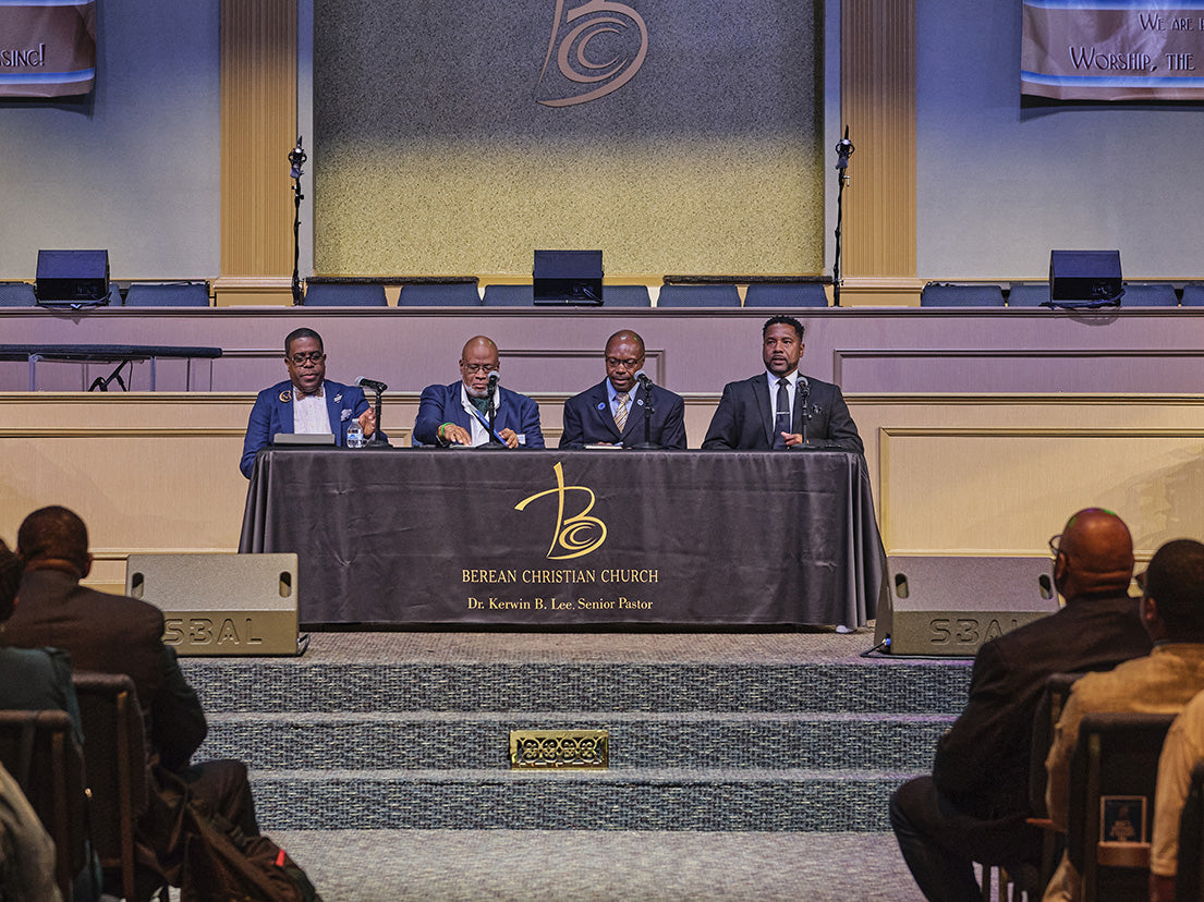 A conference scene with four men seated behind a long table draped in a dark cloth printed with the words 'BERGIAN CHRISTIAN CHURCH' and a gold monogram logo. The stage is lit with warm overhead lights, and several audience members are visible in the foreground.