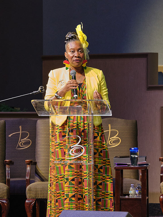 A woman stands behind a clear acrylic podium holding a microphone, wearing a bright yellow blazer and a colorful green, yellow, and red patterned dress with a matching yellow feather headpiece. The setting appears to be a conference or event stage with chairs and a small table in the background.