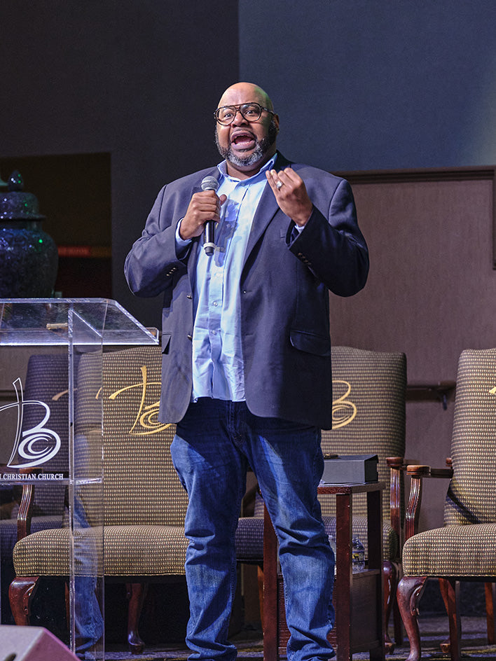 A man wearing glasses, a dark blazer, a light blue shirt, and jeans stands on a stage holding a microphone in front of a pulpit and chairs.