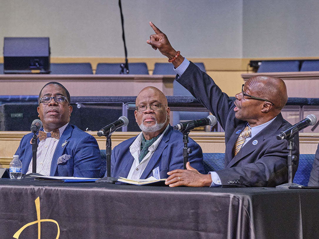 Three men in suits seated at a microphone table in an auditorium or conference hall, with one man raising his hand while speaking. The scene is indoors with rows of seats visible in the background.