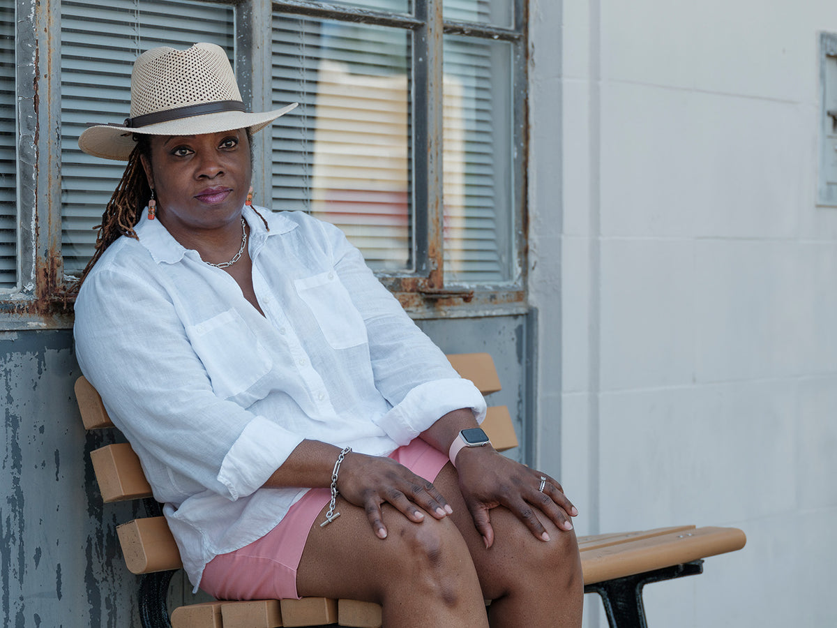 A woman wearing a beige straw hat, white button-up shirt, and pink shorts sits on a wooden bench in front of a weathered window and white wall.
