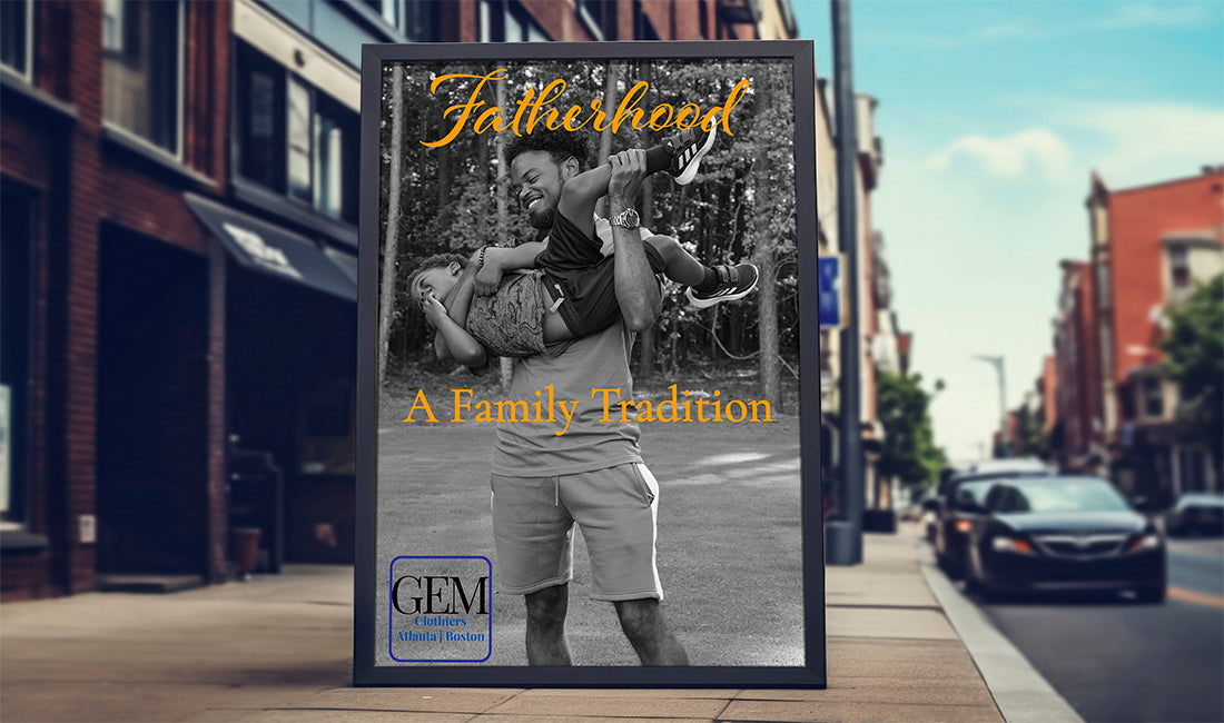 A framed custom family portrait print leaning against a city sidewalk, showing a black-and-white photo of a father carrying his child, with gold script text reading 'Fatherhood' and 'A Family Tradition'. The print has a black frame and a visible 'GEM' logo at the bottom.