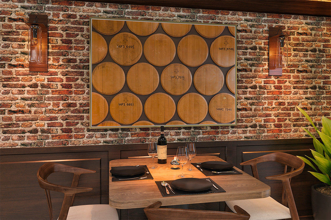 A large framed wall art piece featuring a grid of wooden wine barrels, displayed on a rustic red brick wall above a wooden dining table set with chairs, glasses, and a bottle.
